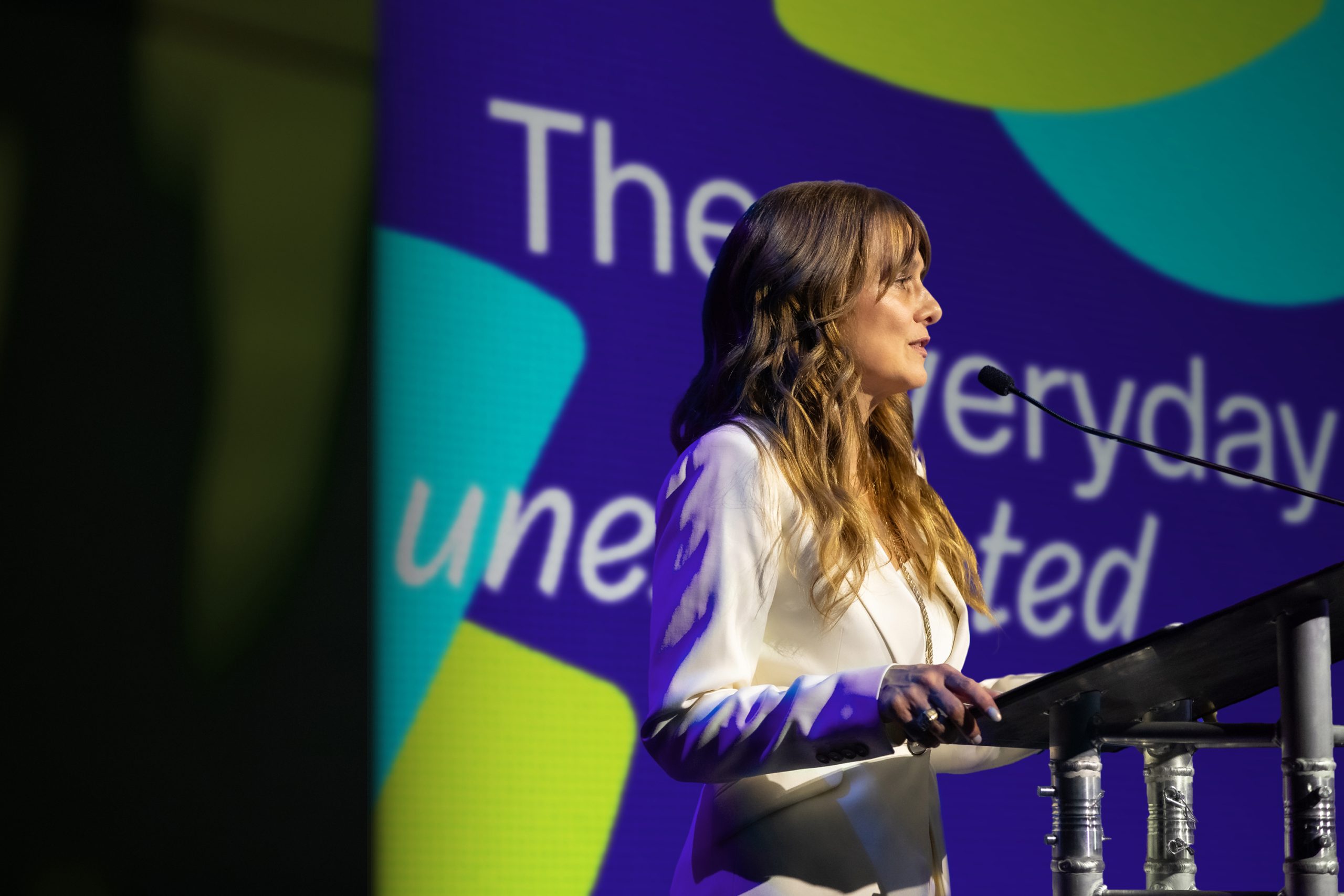Woman speaking at a podium in front of a purple, teal and lime "the everyday unexpected" stage backdrop