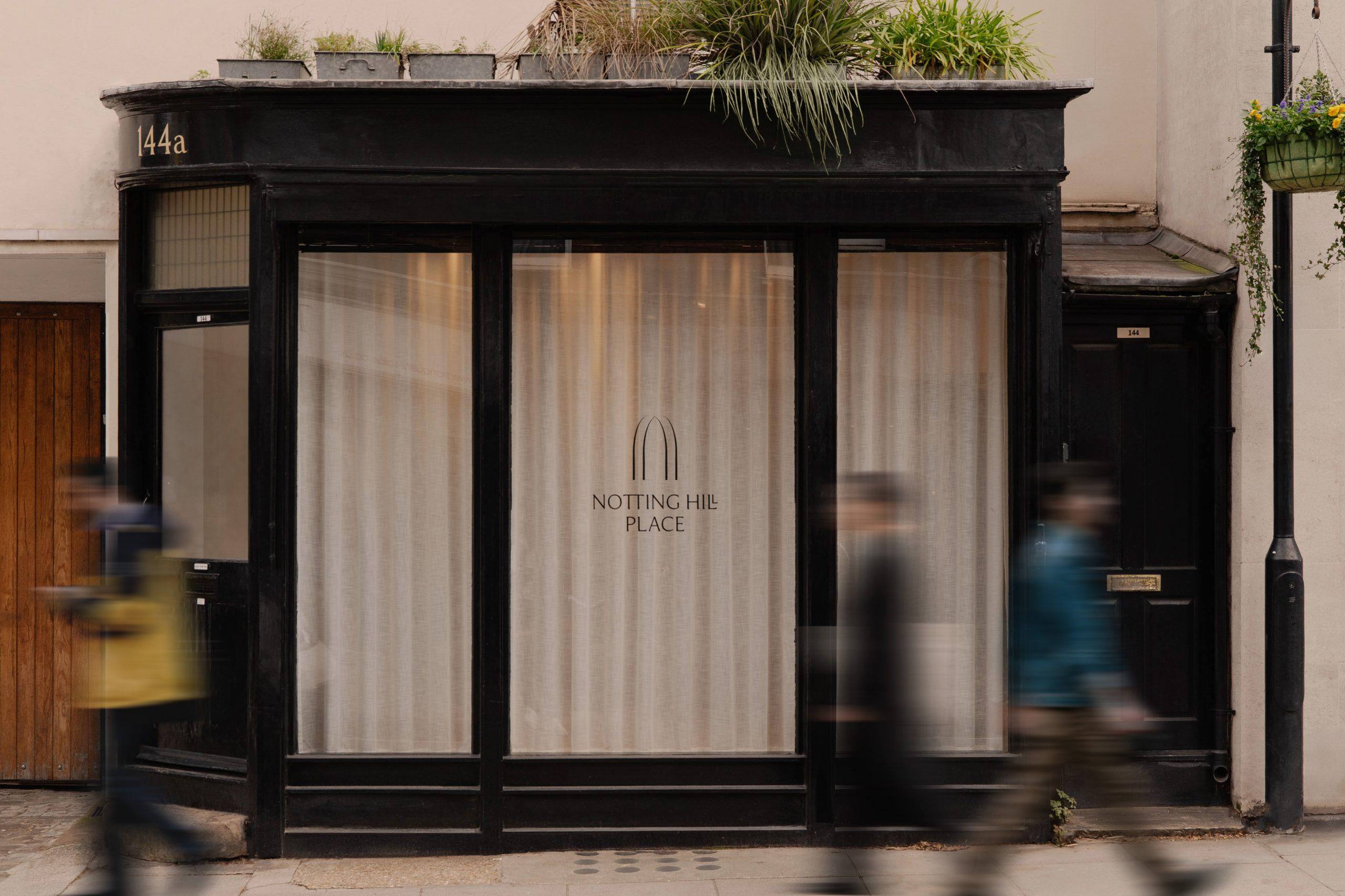 Black-framed Notting Hill Place shopfront at 144a with linen-curtained windows and blurred pedestrians passing by