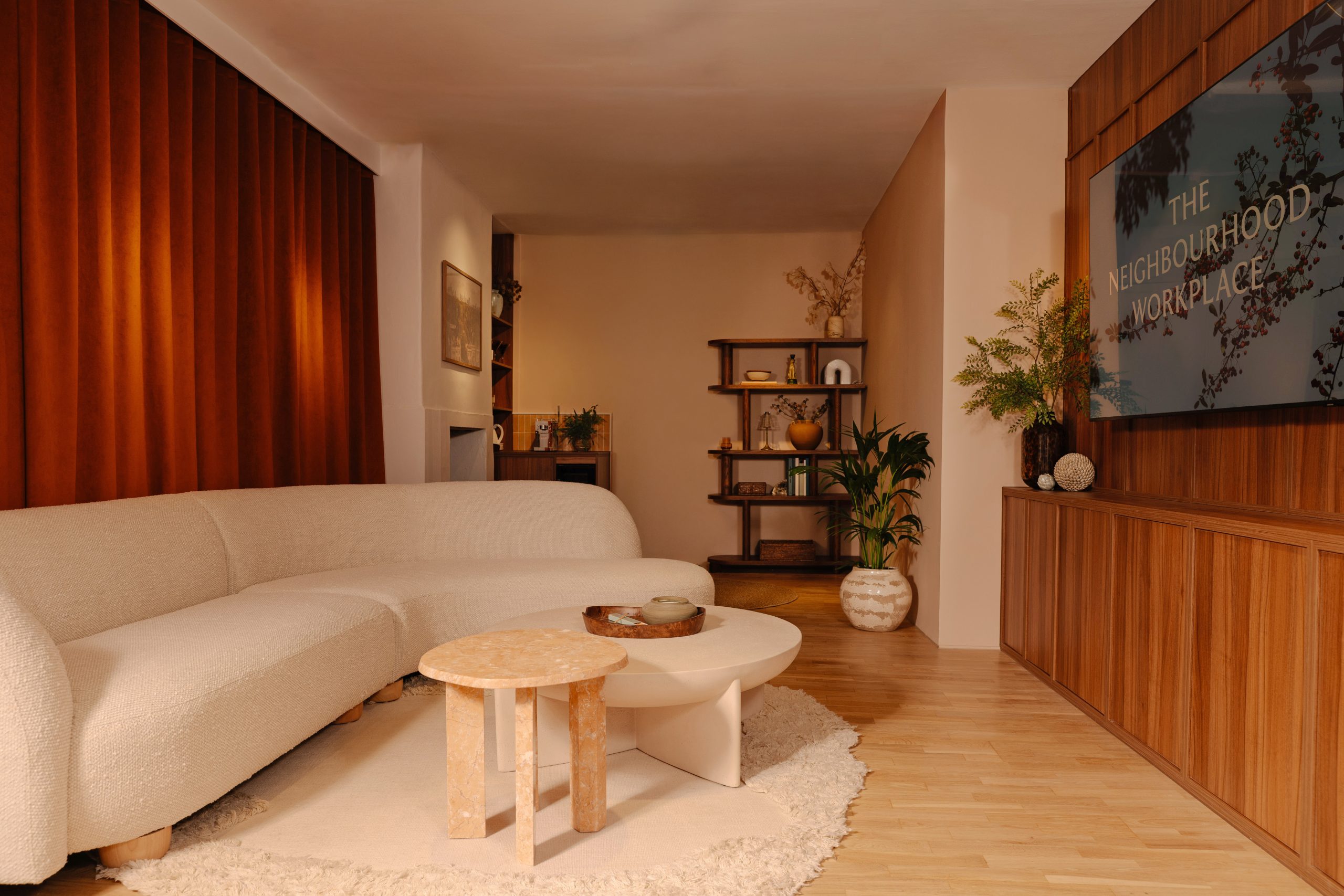Warmly lit lounge interior with a curved bouclé sofa, marble tables, wood paneling, and a screen reading 'The Neighbourhood Workplace'
