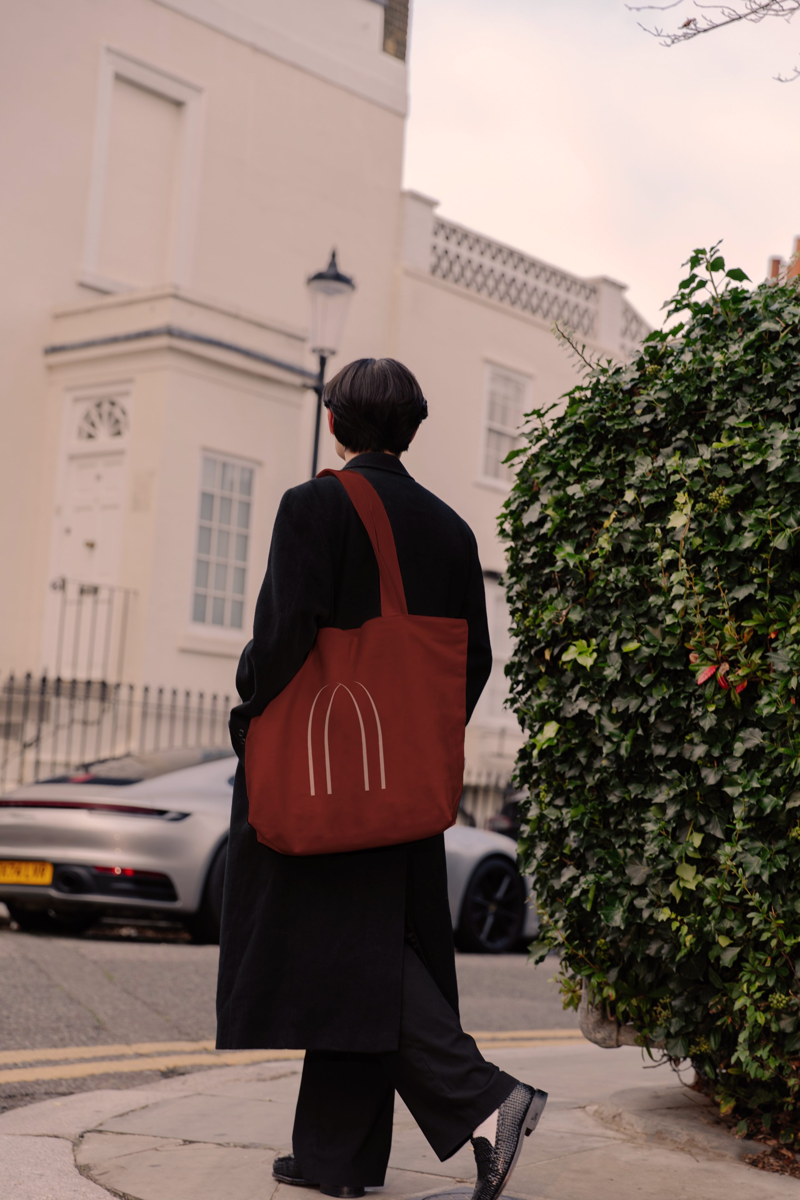 Person in a long black coat walking down a London street carrying a red tote bag with an arch logo