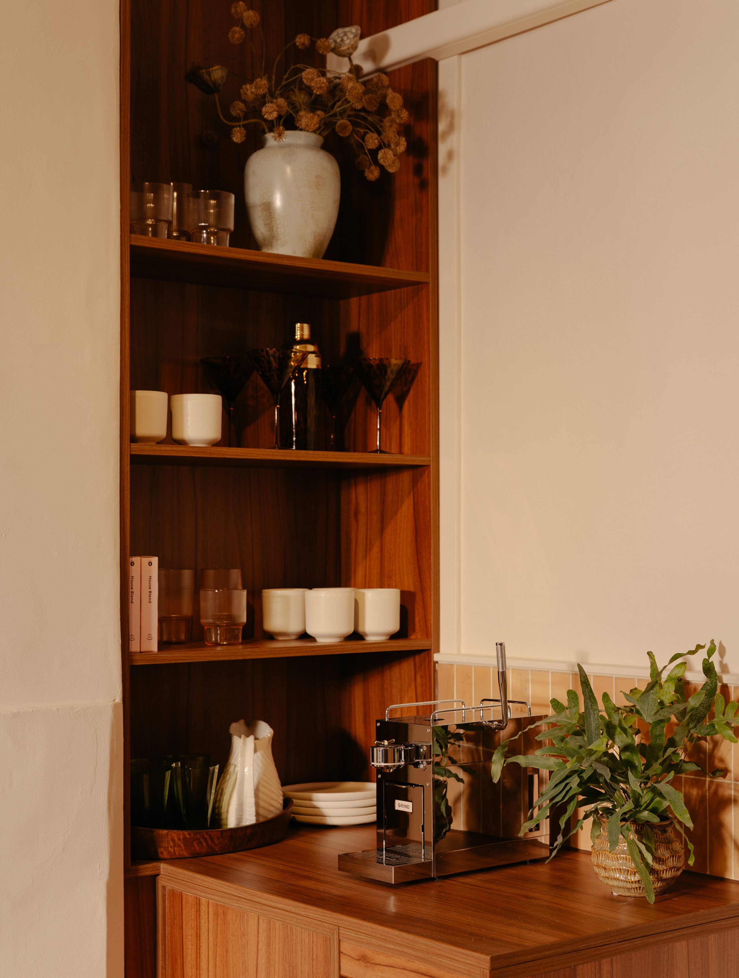 Warm-toned wooden kitchenette with a Grind coffee machine, ceramic cups and glassware on built-in walnut shelves, and a