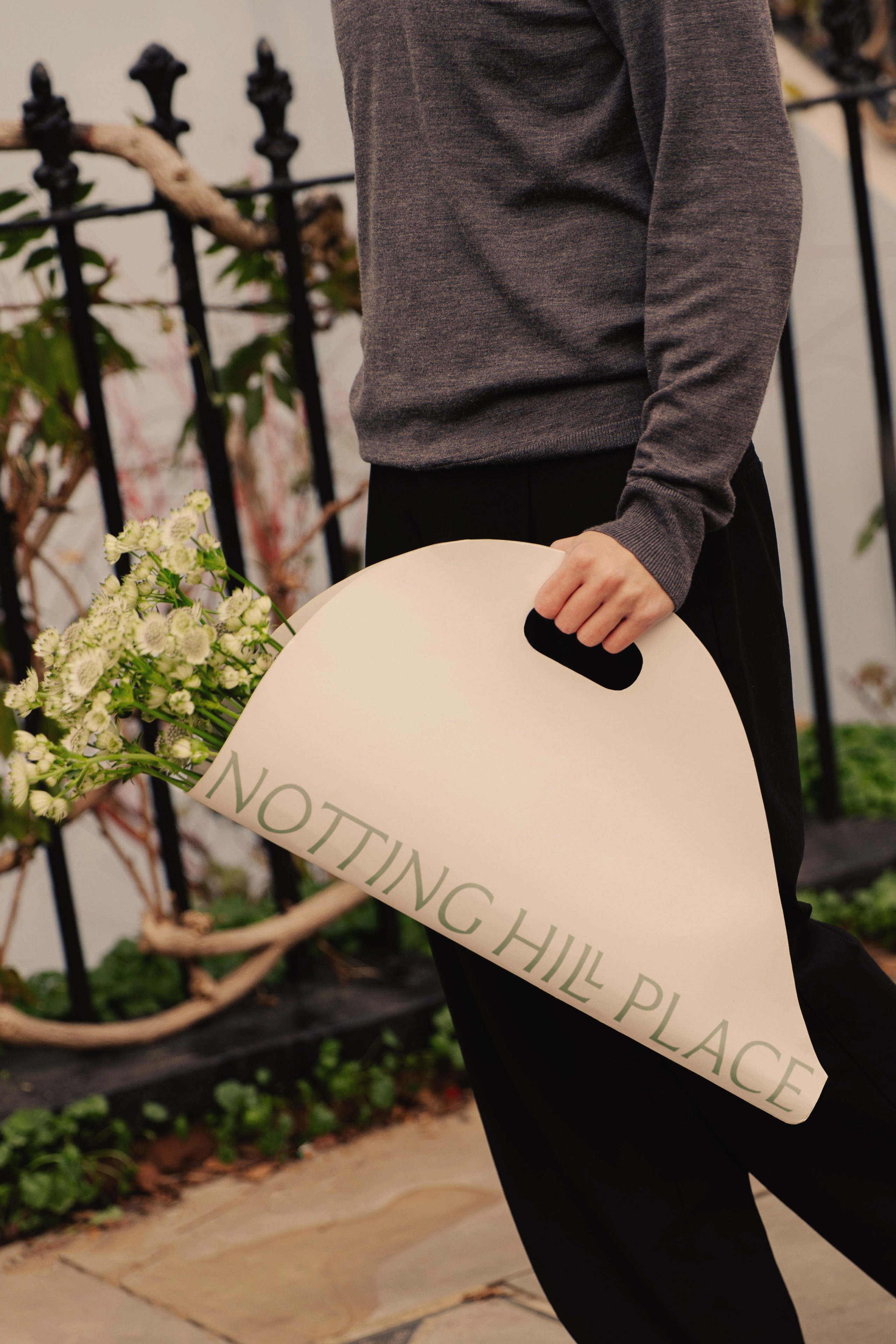 Person carrying a curved Notting Hill Place flower bag with white blooms past a black iron railing