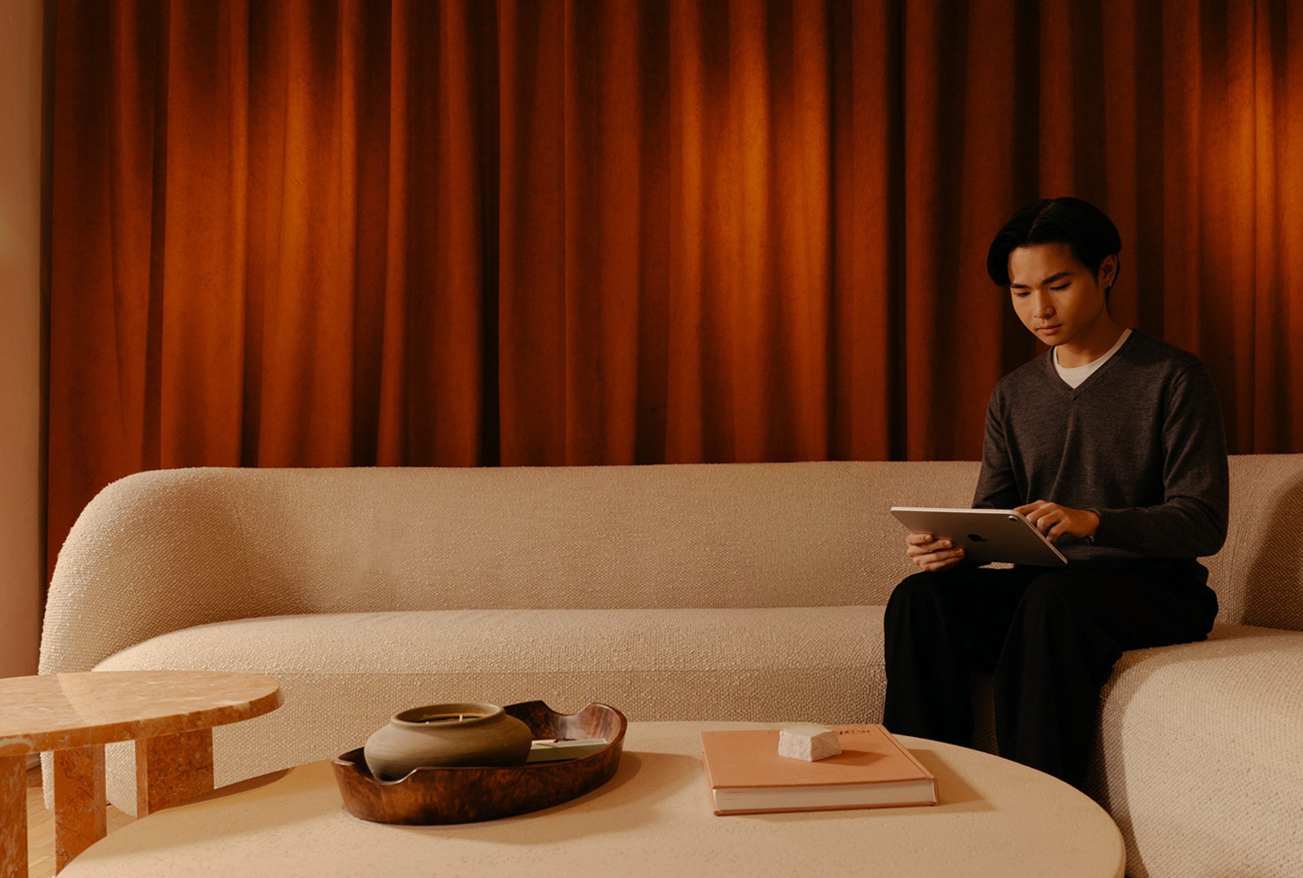 Man in a grey sweater working on a tablet on a curved bouclé sofa in a warmly lit lounge with rust-coloured curtains