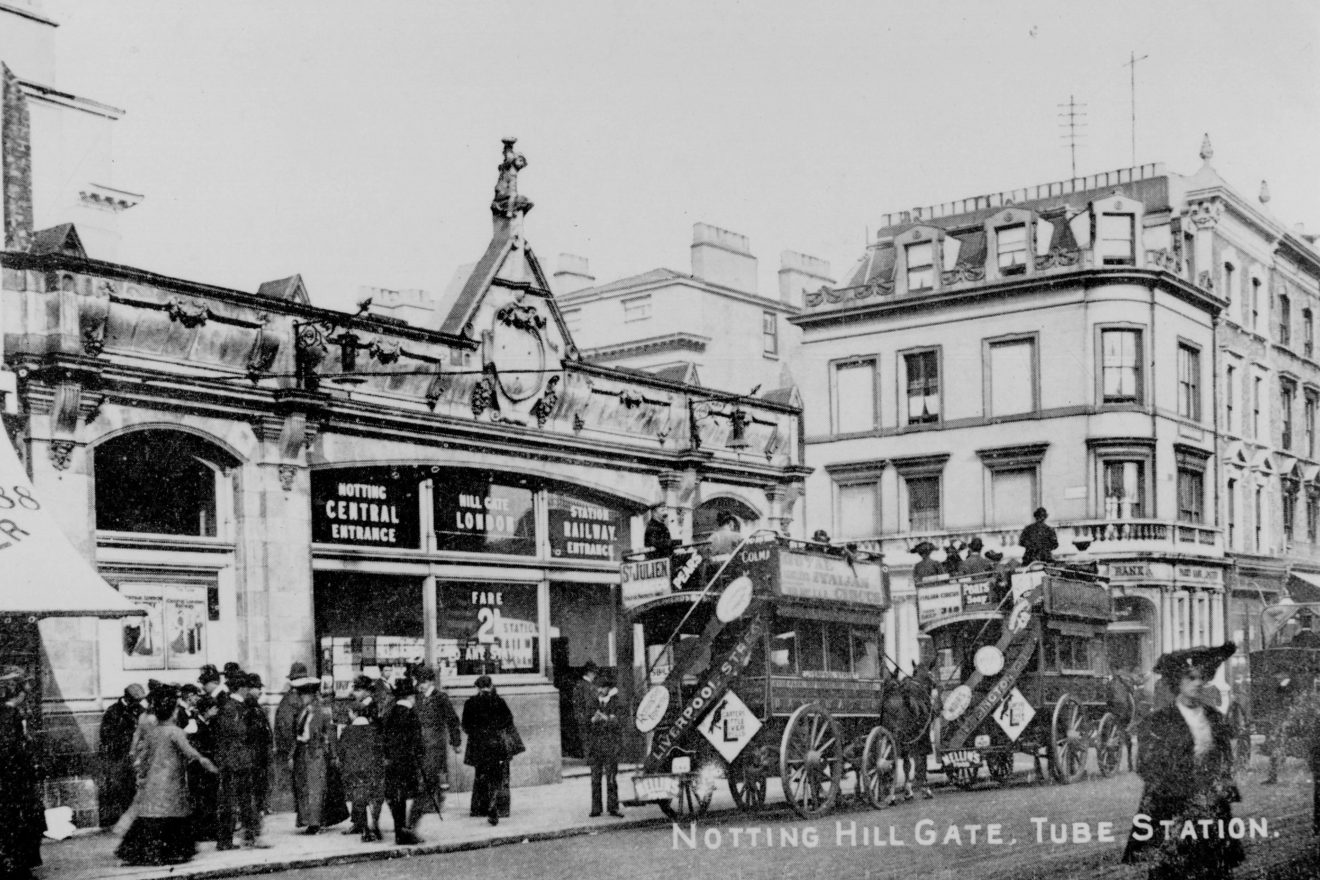 Historic black-and-white photograph of Notting Hill Gate Tube Station with horse-drawn omnibuses and pedestrians in Edwardian dress
