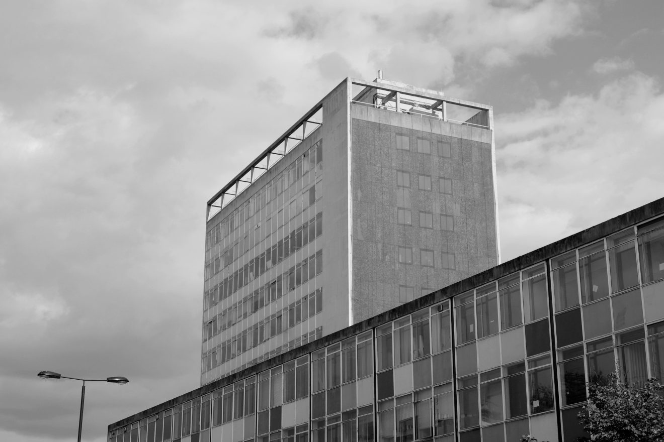 Black-and-white photograph of a mid-century modernist office building with a tall block and lower glass-fronted wing under a cloudy sky